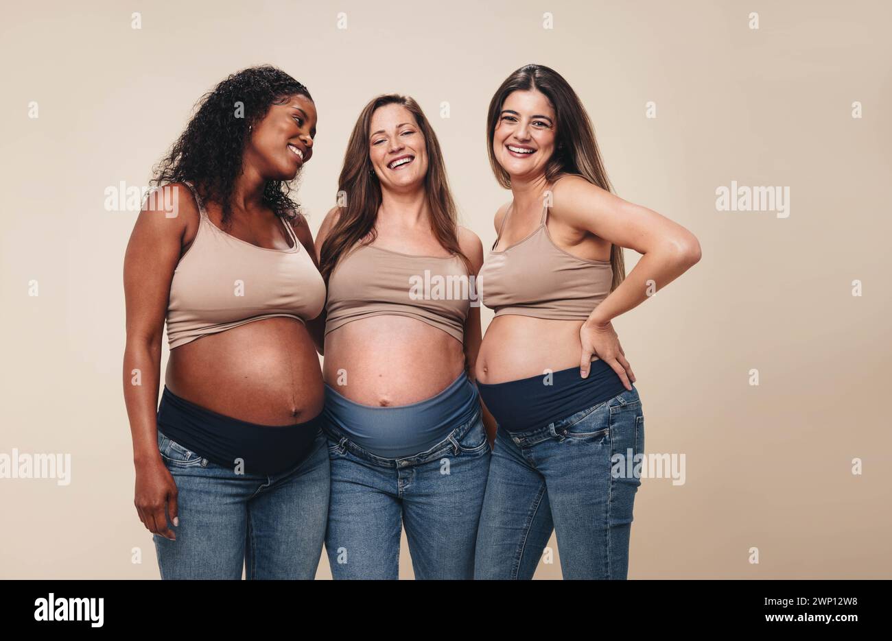 Si aspettano madri con background diversi che si trovino insieme in uno studio, mostrando i loro piccoli dossi. Indossano jeans e reggiseni, sorridendo mentre sostengono Foto Stock