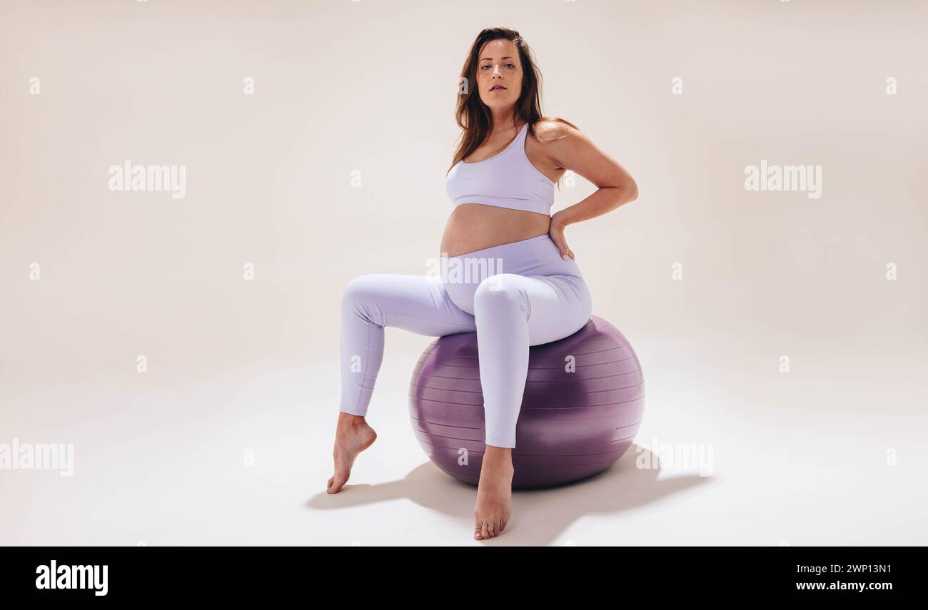 Una giovane donna pratica yoga gravidanza su una palla di stabilità in uno studio. Vestita con abbigliamento fitness, mostra il suo bambino con forza e flexia Foto Stock