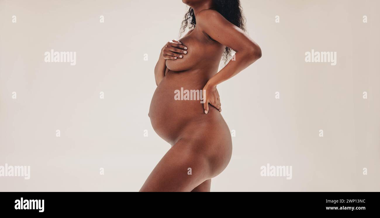 Nello studio, una donna africana sicura di sé nel terzo trimestre di gravidanza mostra con orgoglio il suo bambino bump. Con positività corporea e pelle scura Foto Stock