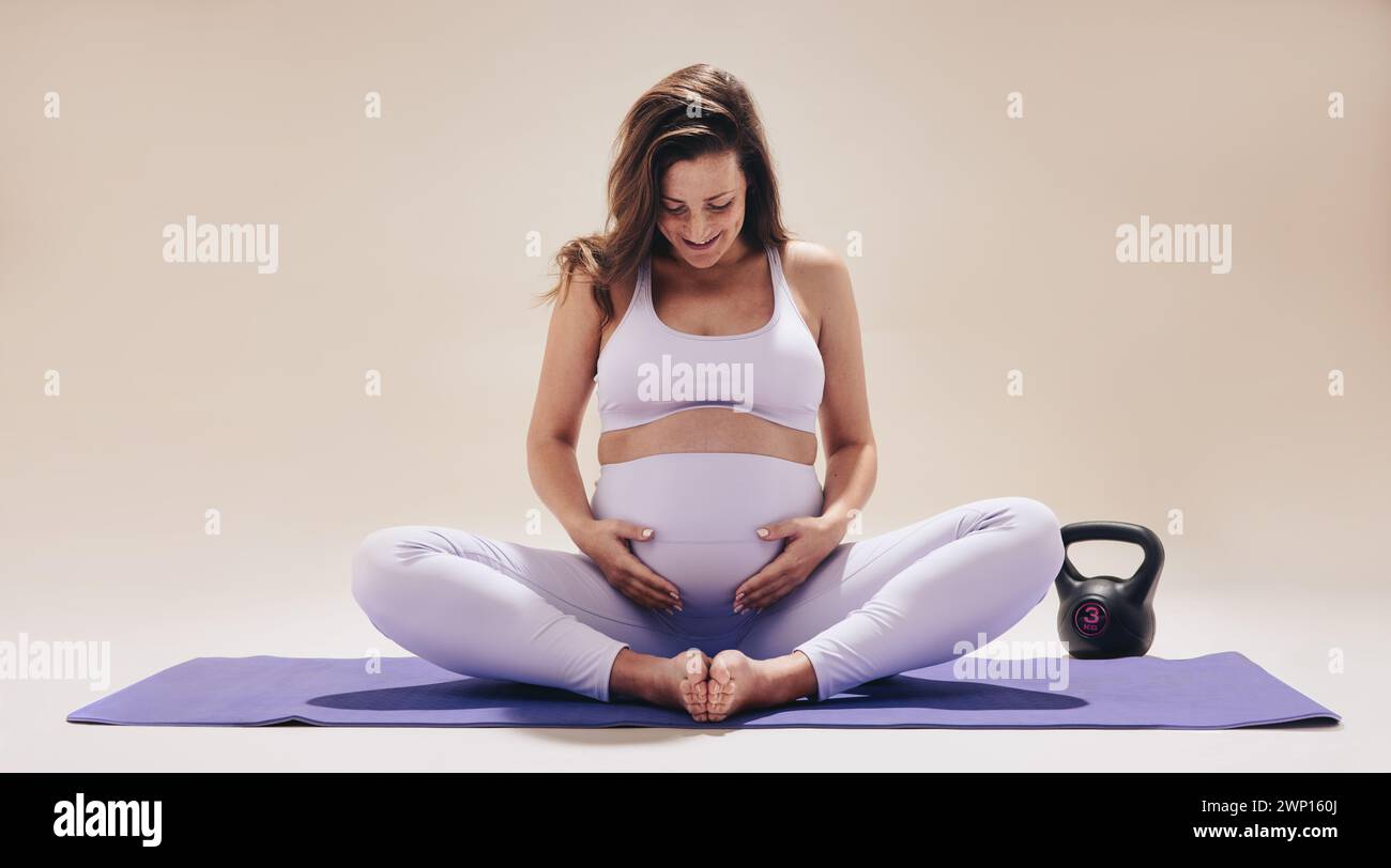 Donna incinta seduta su un tappetino da yoga in uno studio, impegnandosi a uno stile di vita sano nel terzo trimestre. Abbraccia il suo bambino bump, sorridendo Foto Stock