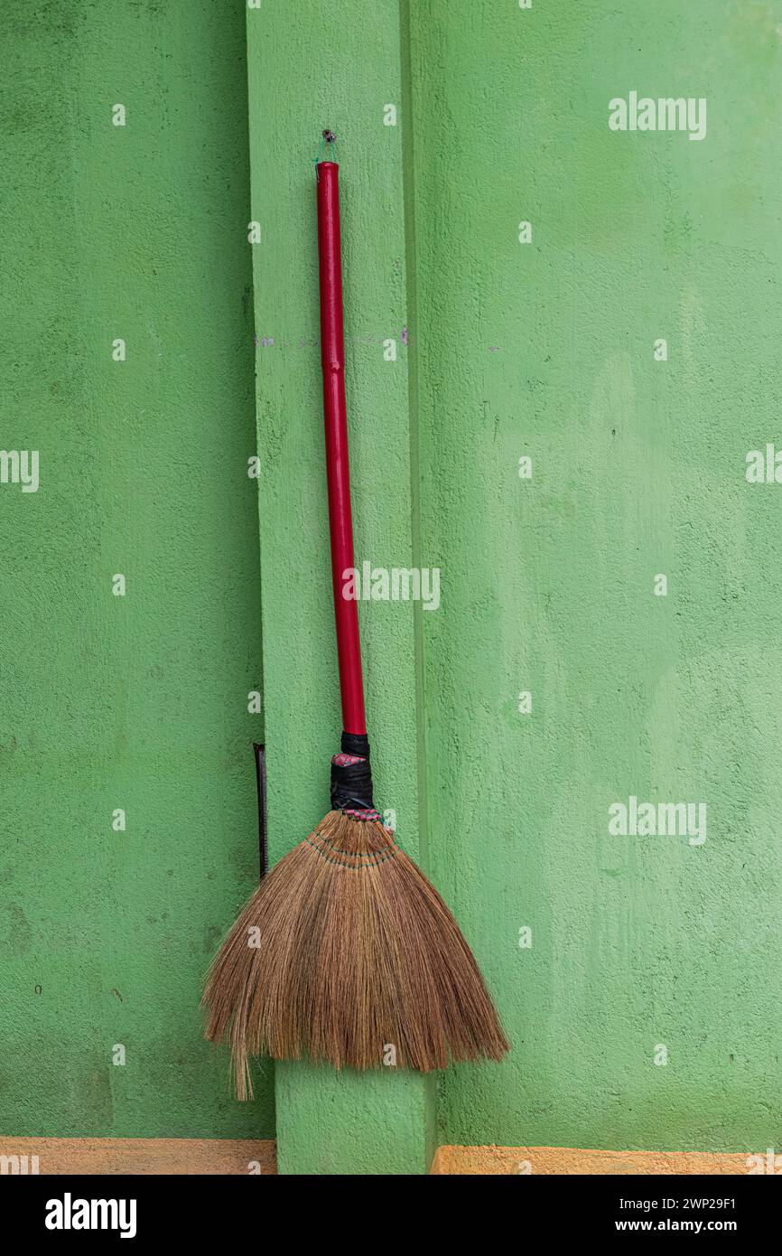 Una semplice scopa in paglia con manico rosso in legno appeso ordinatamente su una parete verde fuori da una casa nel nord rurale del Vietnam. Foto Stock