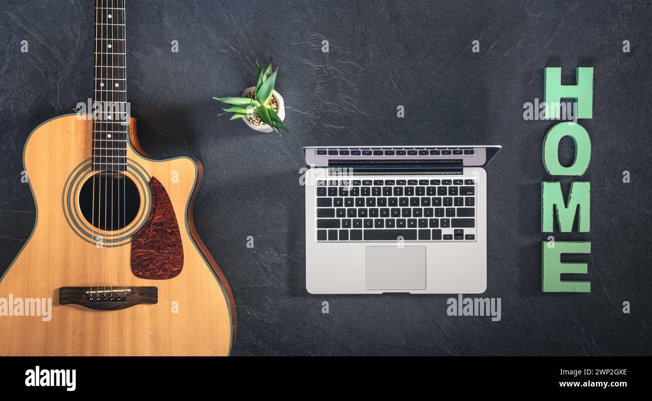 Chitarra acustica, laptop e parola decorativa Home su sfondo scuro, vista dall'alto. Foto Stock
