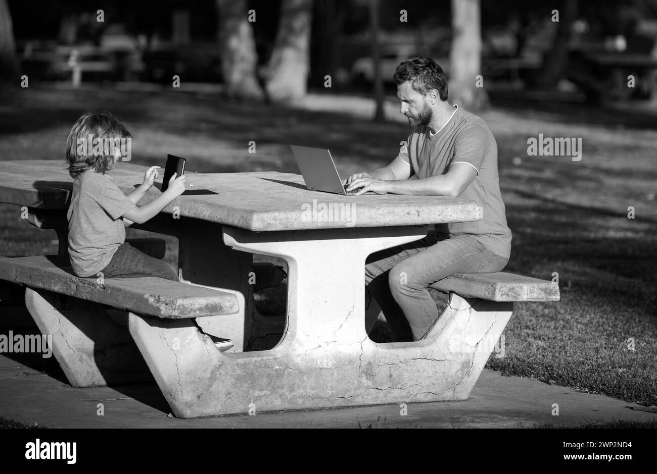 Padre insegna al figlio a usare il laptop, il papà e il bambino della scuola guardando lo schermo del computer e il tablet, giocando, guardando il video, seduto sull'erba Foto Stock