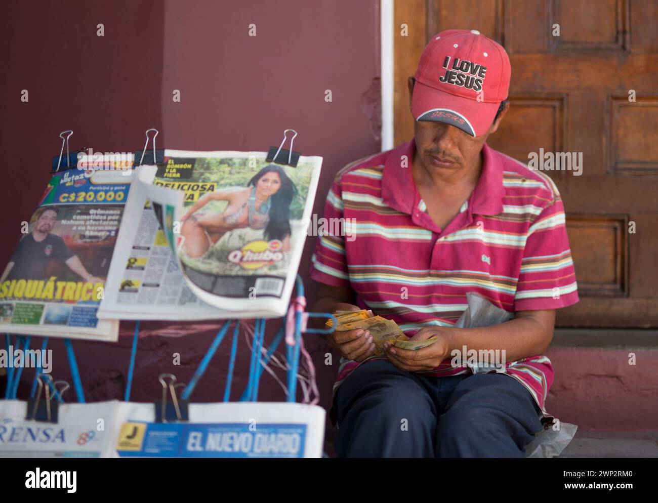 25/02/16 Un venditore di giornali, che indossa un cappello "i Love Jesus", conta i soldi come una grande donna al petto, è esposto su uno dei suoi giornali a Granada, Foto Stock