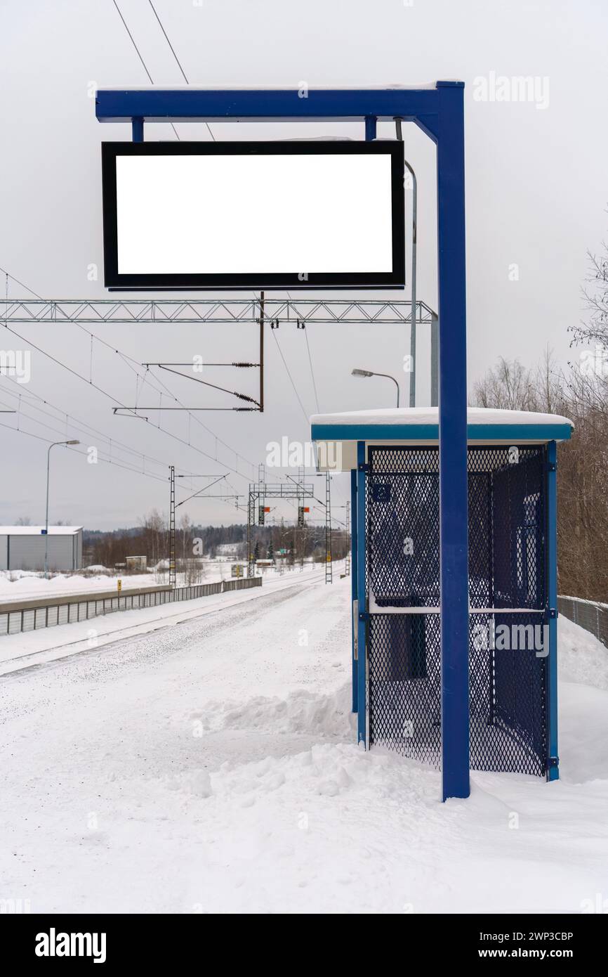 Piattaforma vuota della stazione ferroviaria in una giornata di neve, con un rifugio blu e un cartellone vuoto con pista di ritaglio. Foto Stock
