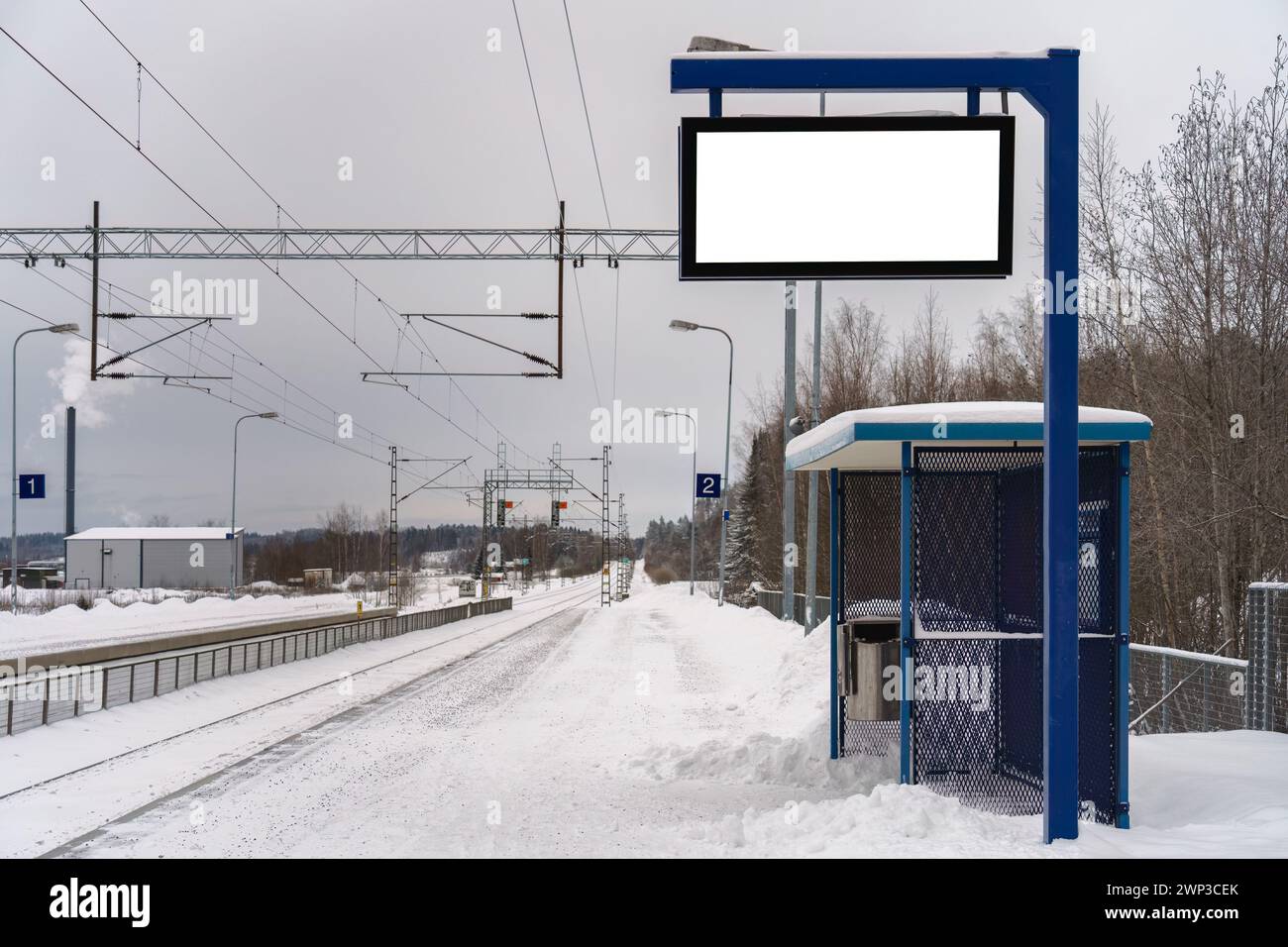 Piattaforma vuota della stazione ferroviaria in una giornata di neve, con un rifugio blu e un cartellone vuoto con pista di ritaglio. Foto Stock