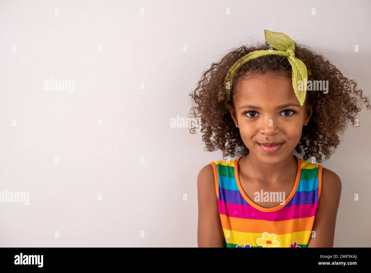 Giovane ragazza nera con capelli ricci, con canotta a righe arcobaleno e fascia verde, isolata su sfondo bianco, felice e sorridente. an minimalista Foto Stock