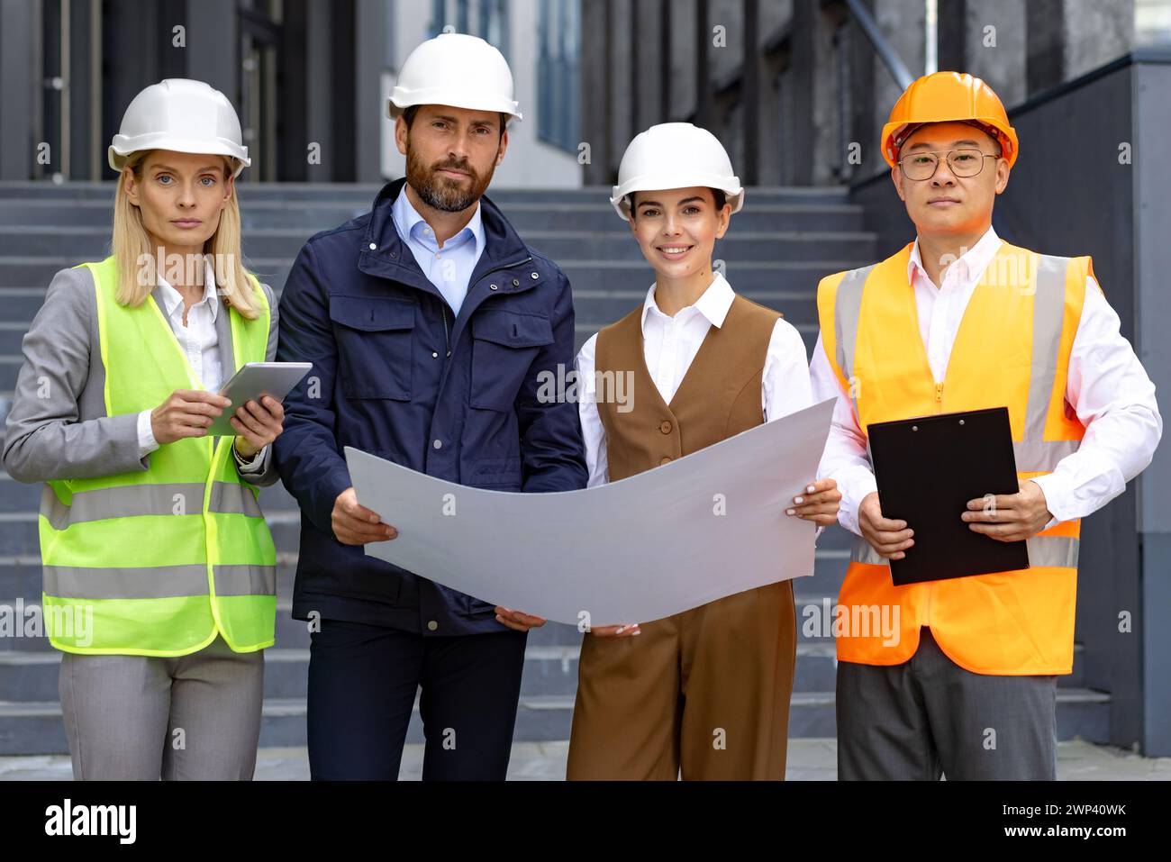 Quattro professionisti del settore edile in Hard Hats che esaminano il progetto all'esterno di un edificio moderno, concentrati e determinati. Foto Stock