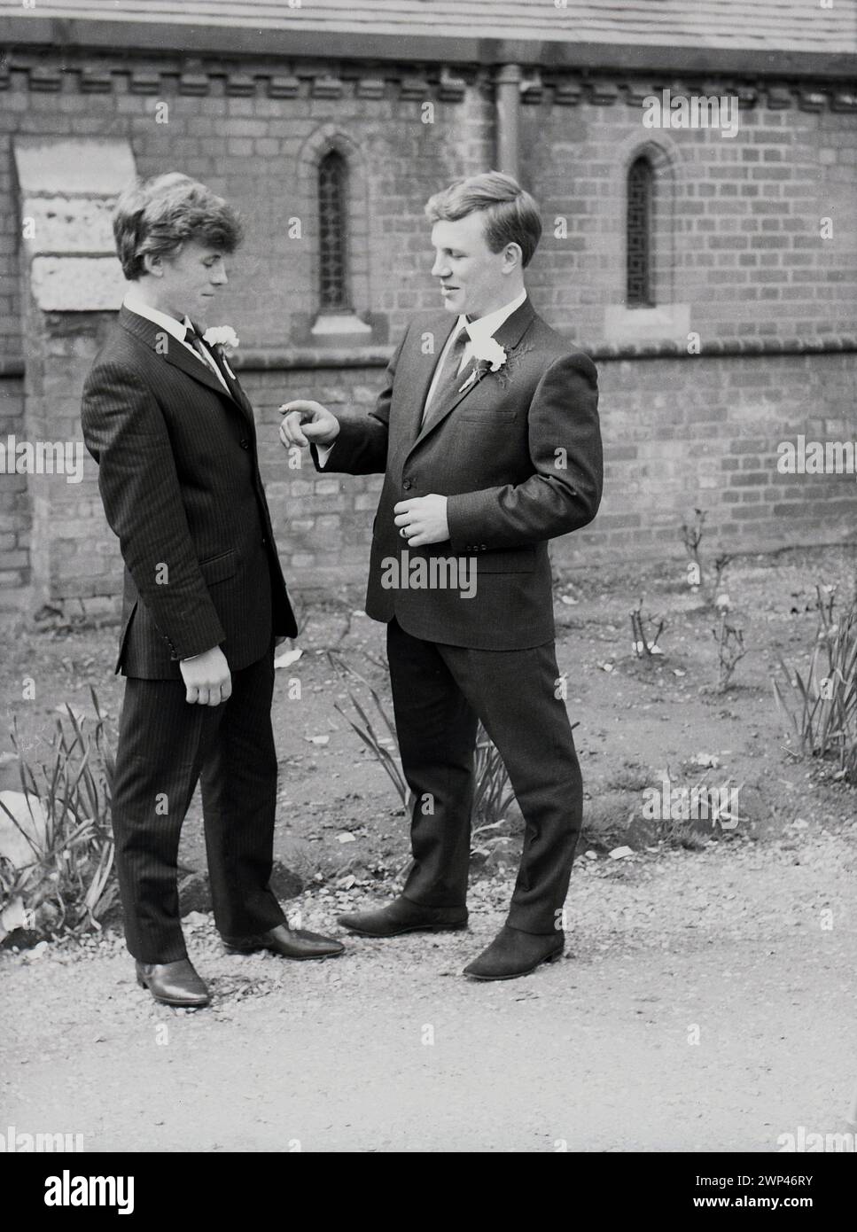 Anni '1960, storici, due giovani uomini, di piccola statura, insieme nei loro migliori abiti e stivali a punta, che partecipano a un matrimonio, in piedi a parlare su un sentiero fuori dalla chiesa, Inghilterra, Regno Unito. Foto Stock