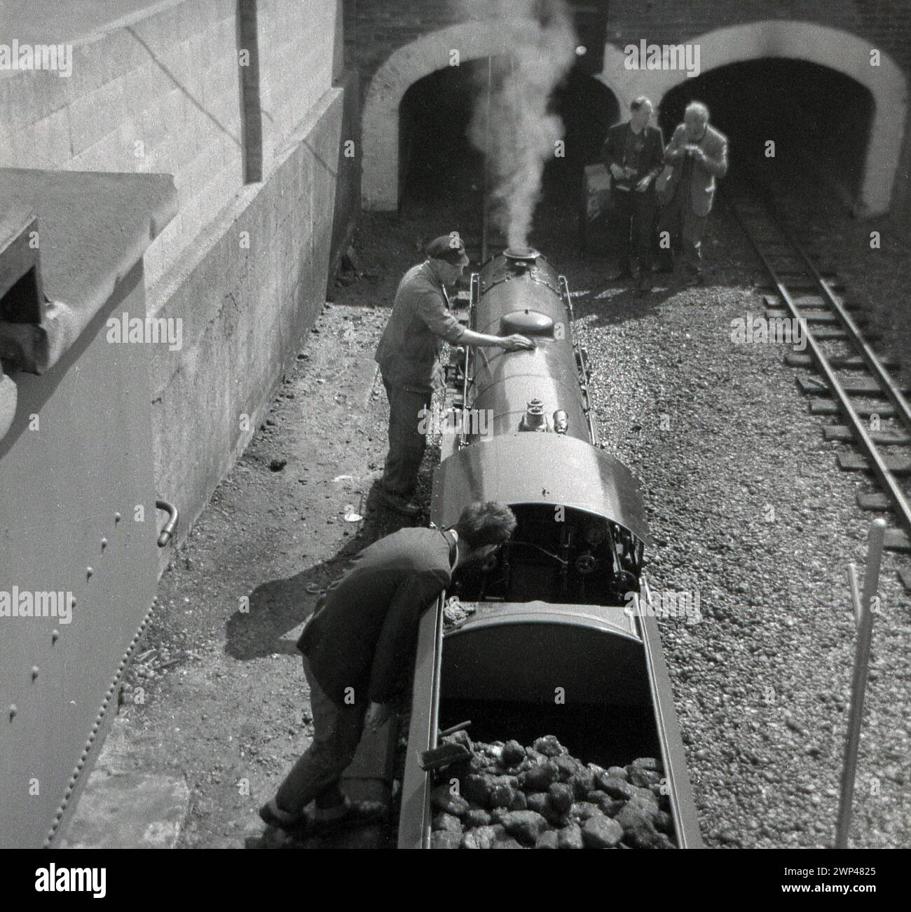 1960, storico, una vista dall'alto di diversi appassionati di treni maschi accanto a una locomotiva a vapore in miniatura, con trasporto di carbone, su un binario ferroviario sommerso fuori da un doppio tunnel, Inghilterra, Regno Unito. Foto Stock