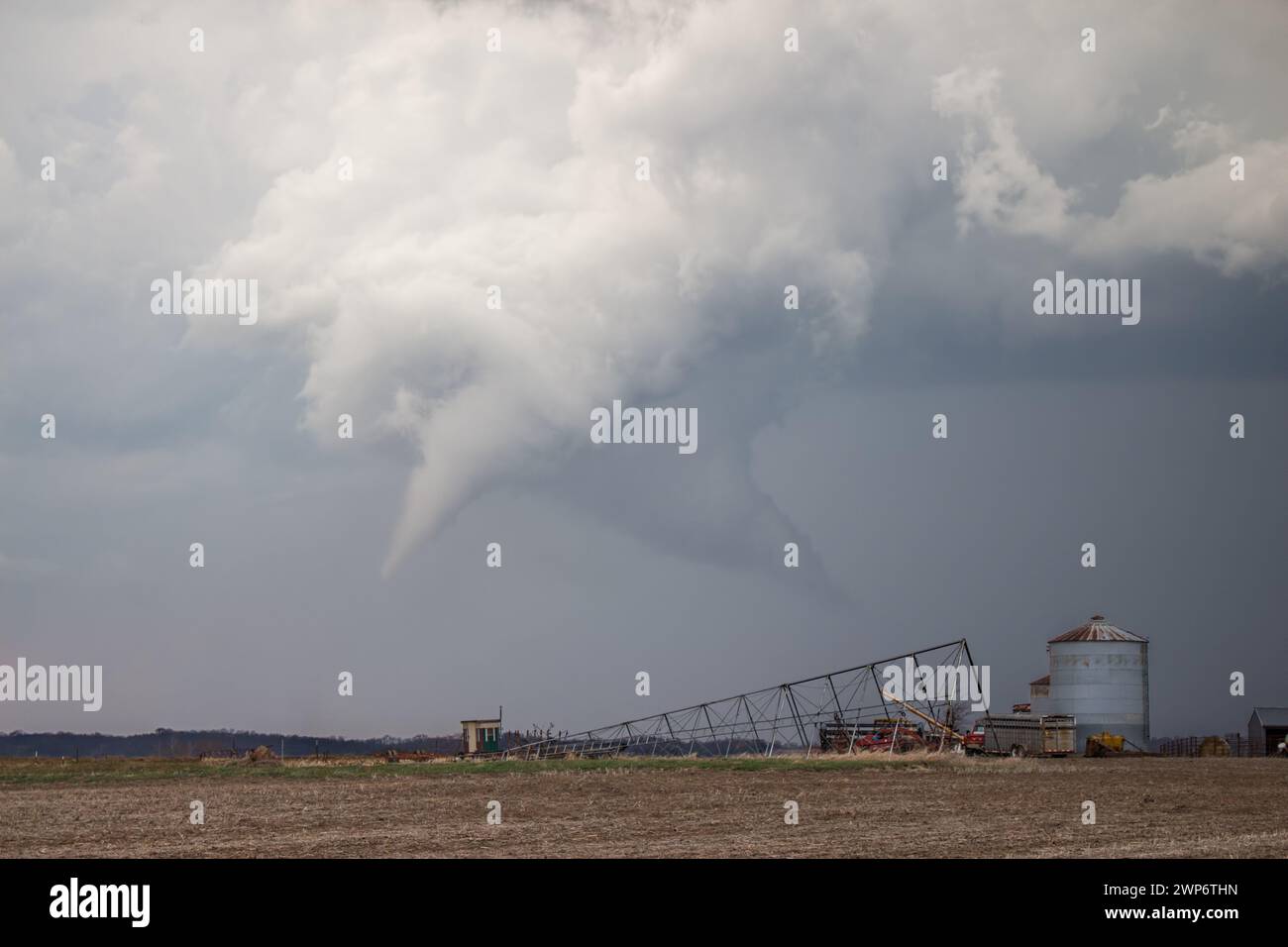 Un tornado a cono bianco è appeso sotto una nuvola di tempesta su terreni agricoli rurali con edifici agricoli e attrezzature in primo piano. Foto Stock