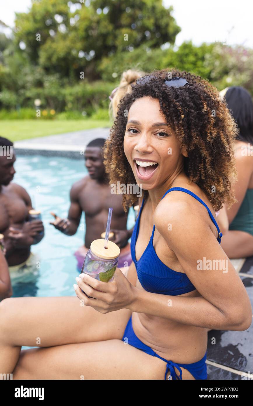 Una donna birazziale in costume blu si gode una festa in piscina, sorseggiando un drink Foto Stock