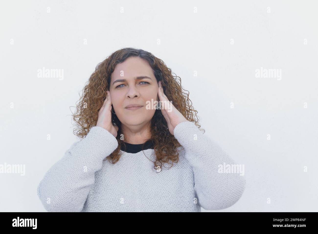 giovane donna attraente con lunghi capelli ricci e occhi blu copre le orecchie con le mani, con sfondo bianco e spazio di copia Foto Stock