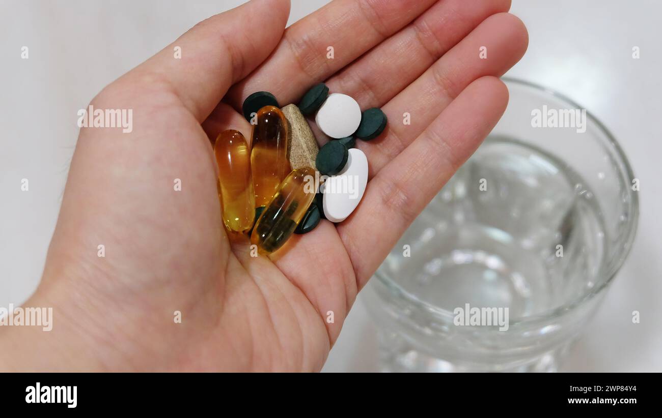 Mani che tengono pillole e capsule assortite di integratori. Con un bicchiere d'acqua sullo sfondo. Foto Stock