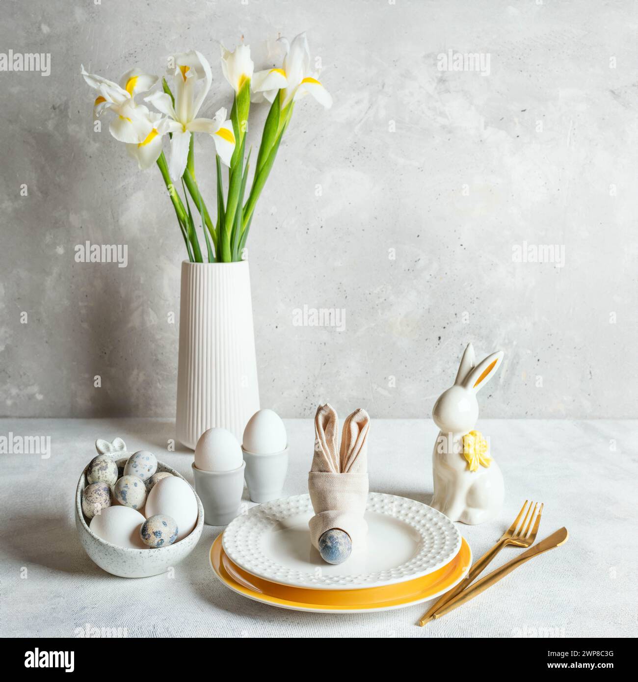 Tavolo pasquale bianco monocromatico. Uova dai colori naturali, piatti figurati, coniglietto in ceramica, tovagliolo come muso coniglietto, bouquet di fiori bianchi di Iris sul tavolo. Poliziotto Foto Stock