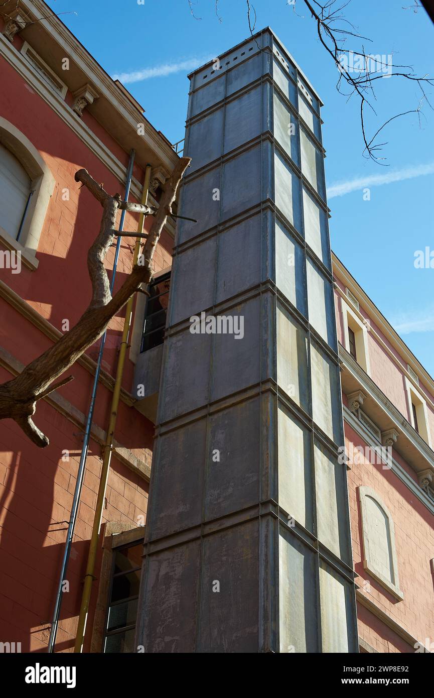 Ascensore in metallo grigio collegato a un edificio rosso, utilizzato per lavori di ristrutturazione e costruzione, catturato in una giornata di sole. Foto Stock