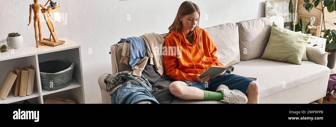 una ragazza adolescente che legge un libro mentre sta seduta su un divano disordinato accanto a una pila di vestiti nell'appartamento, uno striscione Foto Stock