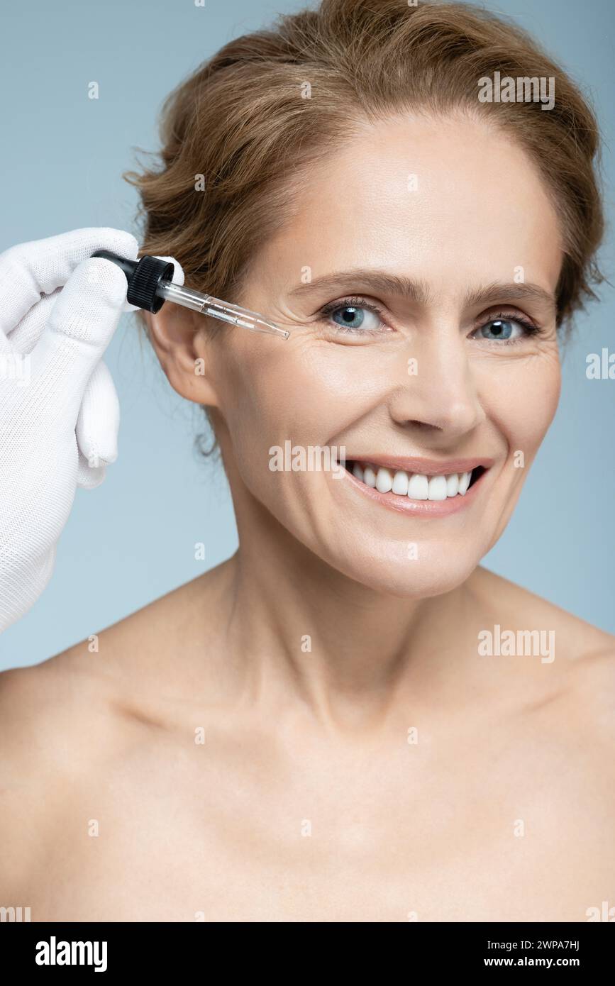 cura della pelle del viso con olio cosmetico e siero per il trattamento. Bella donna caucasica che applica siero sul viso, isolata su sfondo blu Foto Stock