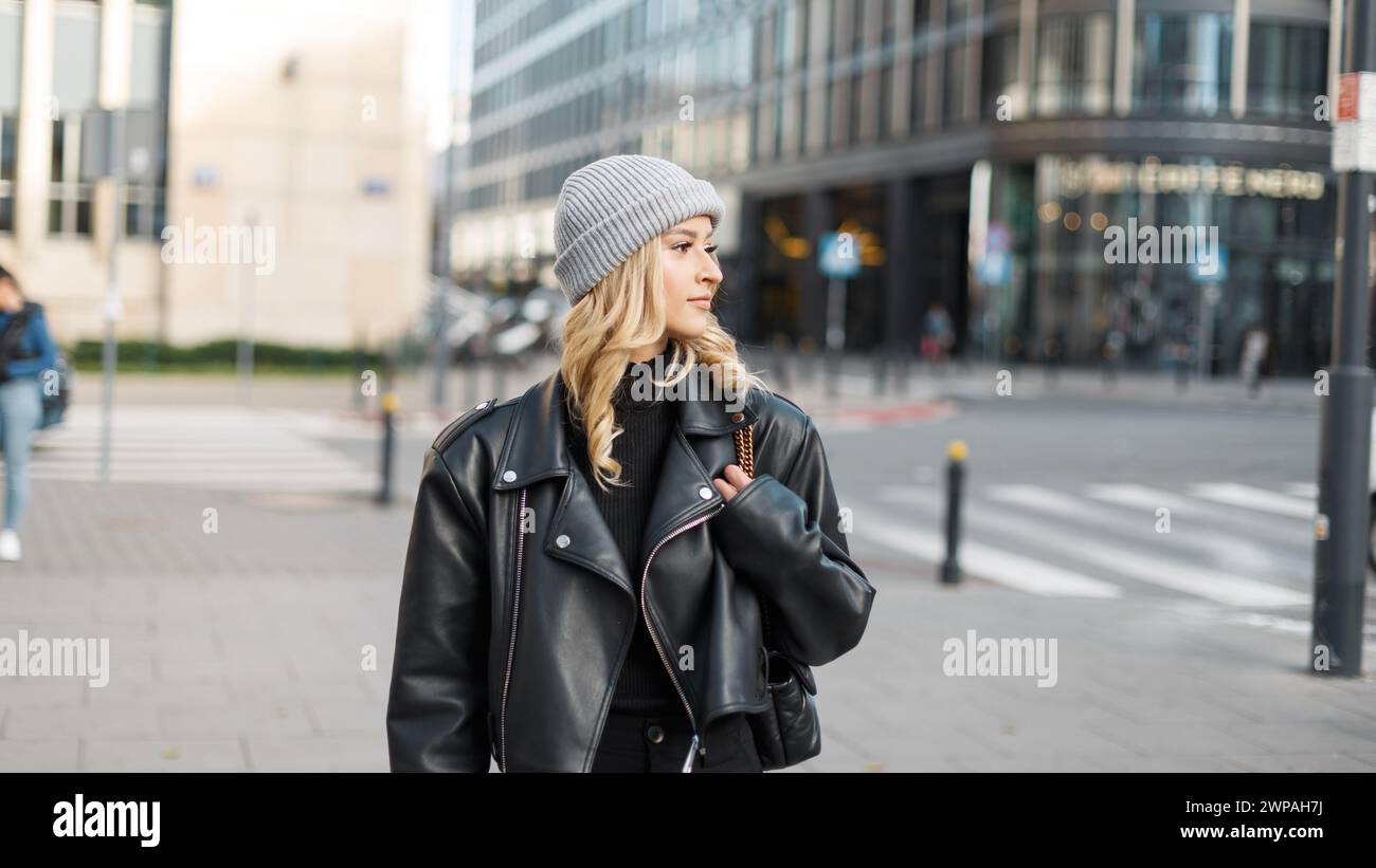 Elegante modella urbana da donna giovane in abiti alla moda con Un cappello in maglia e una giacca in pelle che cammina per strada Foto Stock