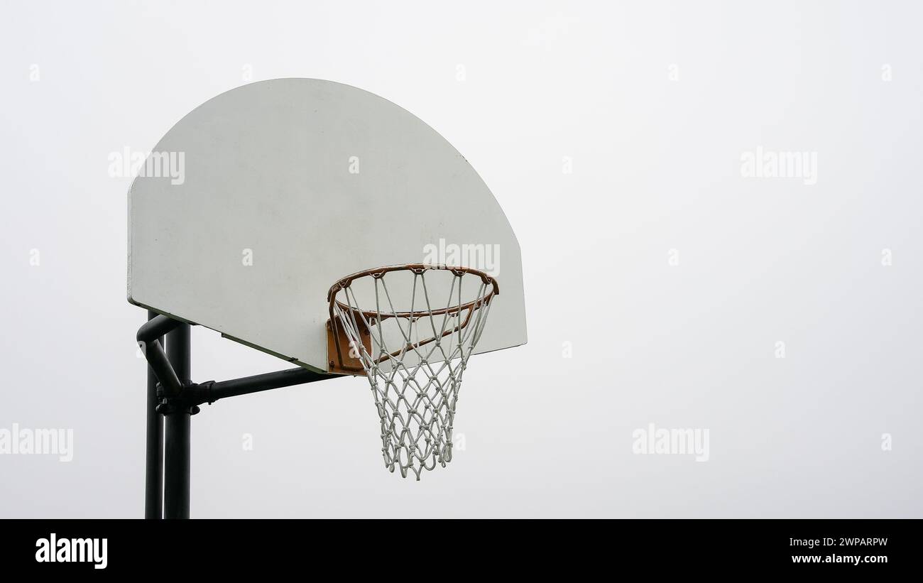 Un unico canestro da basket si erge alto con il suo tabellone bianco e il bordo arancione, adagiato contro un cielo limpido e pallido, in attesa che i giocatori inizino una partita. Foto Stock