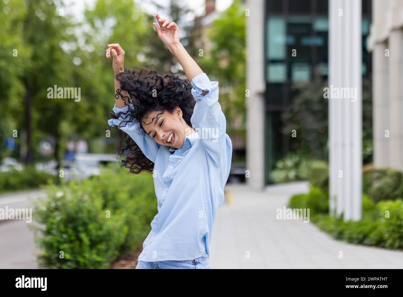 Una giovane donna esuberante con i capelli ricci danzano gioiosamente su un marciapiede urbano. La sua espressione vibrante e il suo movimento spensierato catturano un senso di libertà e felicità nell'ambiente cittadino. Foto Stock