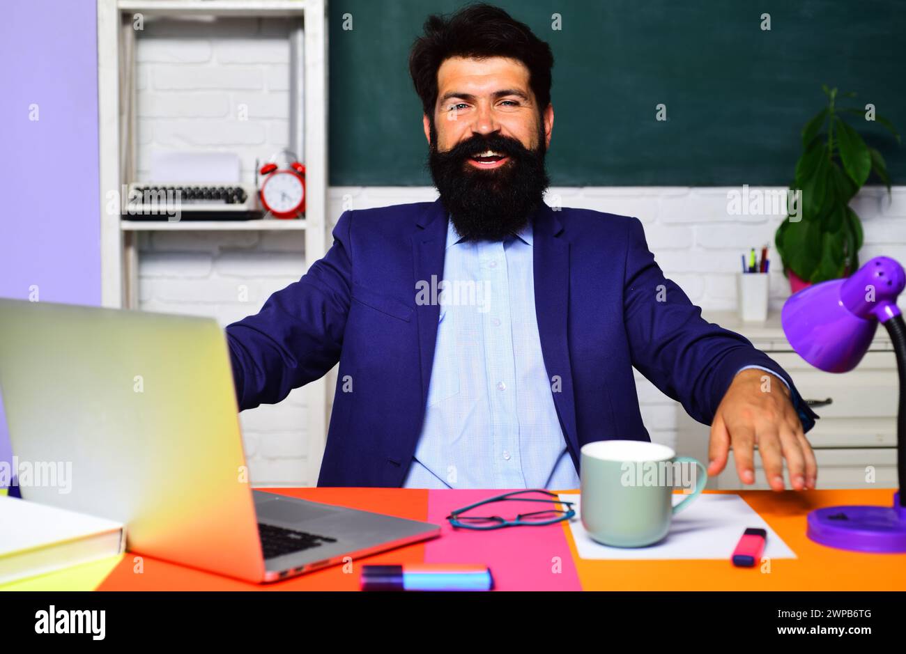 Un insegnante sorridente o uno studente che utilizza un laptop seduto alla scrivania e studia online. Corsi di formazione virtuali. Insegnante di sesso maschile in preparazione per test o esami in Foto Stock