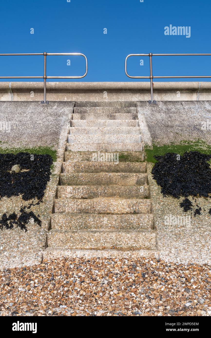 Gradini sul muro del mare che conducono dalla spiaggia di ciottoli alla strada di Hill Head vicino a Titchfield, Hampshire, Inghilterra, Regno Unito, con alghe e cielo blu Foto Stock