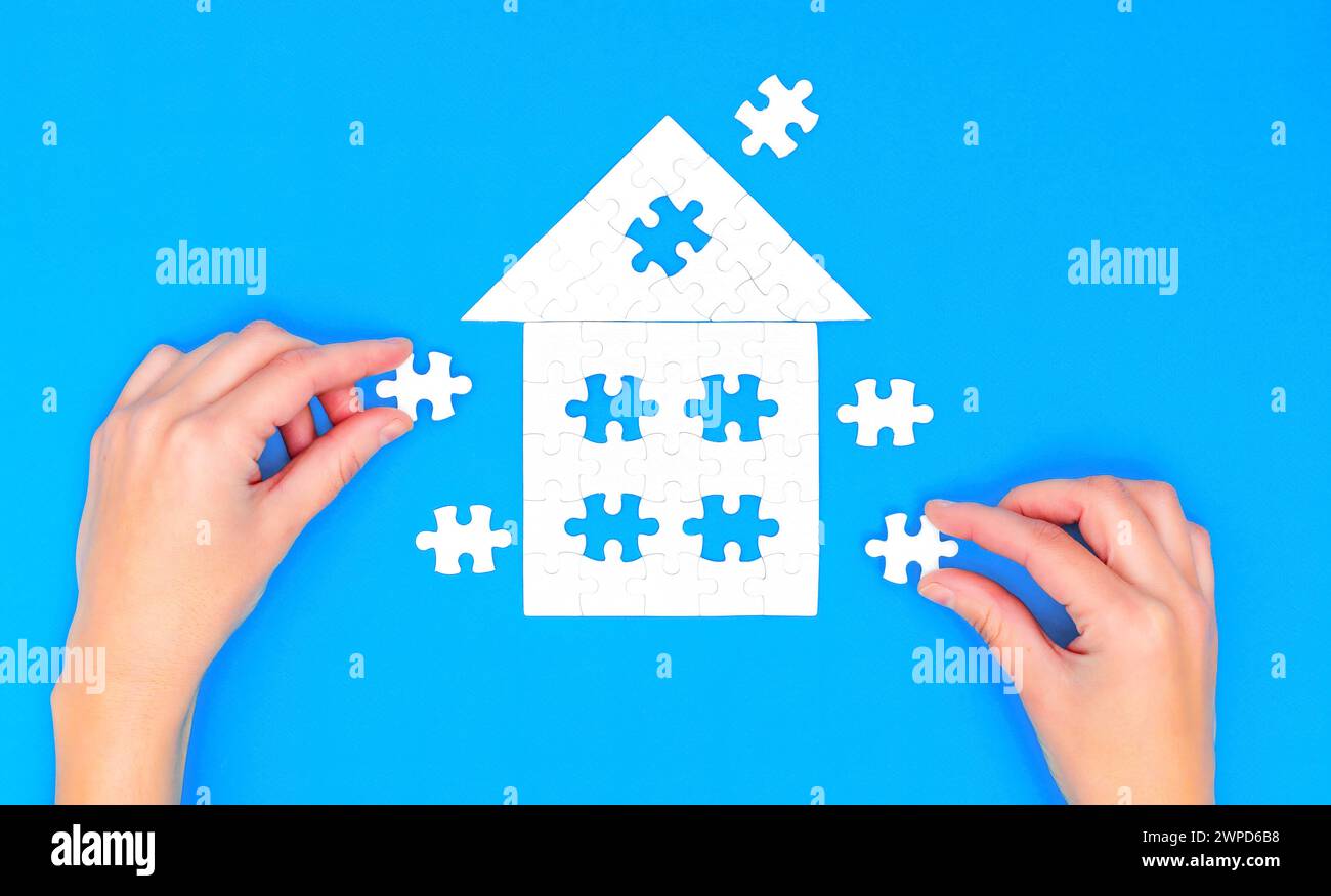 Un primo piano di mani femminili che combinano un puzzle a forma di casa con un tetto all'anca, su uno sfondo blu rilassante. Foto Stock