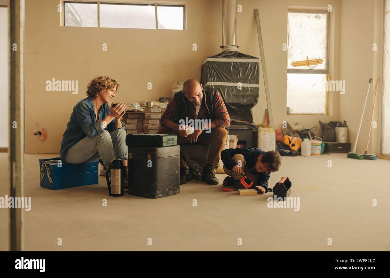 I genitori riposano durante il loro progetto di ristrutturazione della casa, gustando un caffè insieme mentre il loro bambino gioca con gli strumenti nelle vicinanze. Il loro lavoro di squadra brilla come Foto Stock