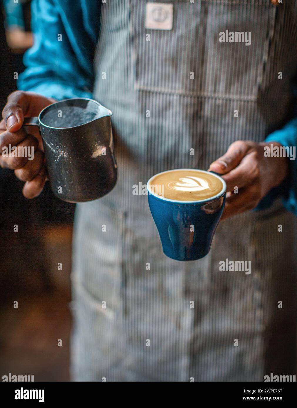 barista con grembiule che versa il latte montato nella tazza di caffè creando un bianco piatto Foto Stock