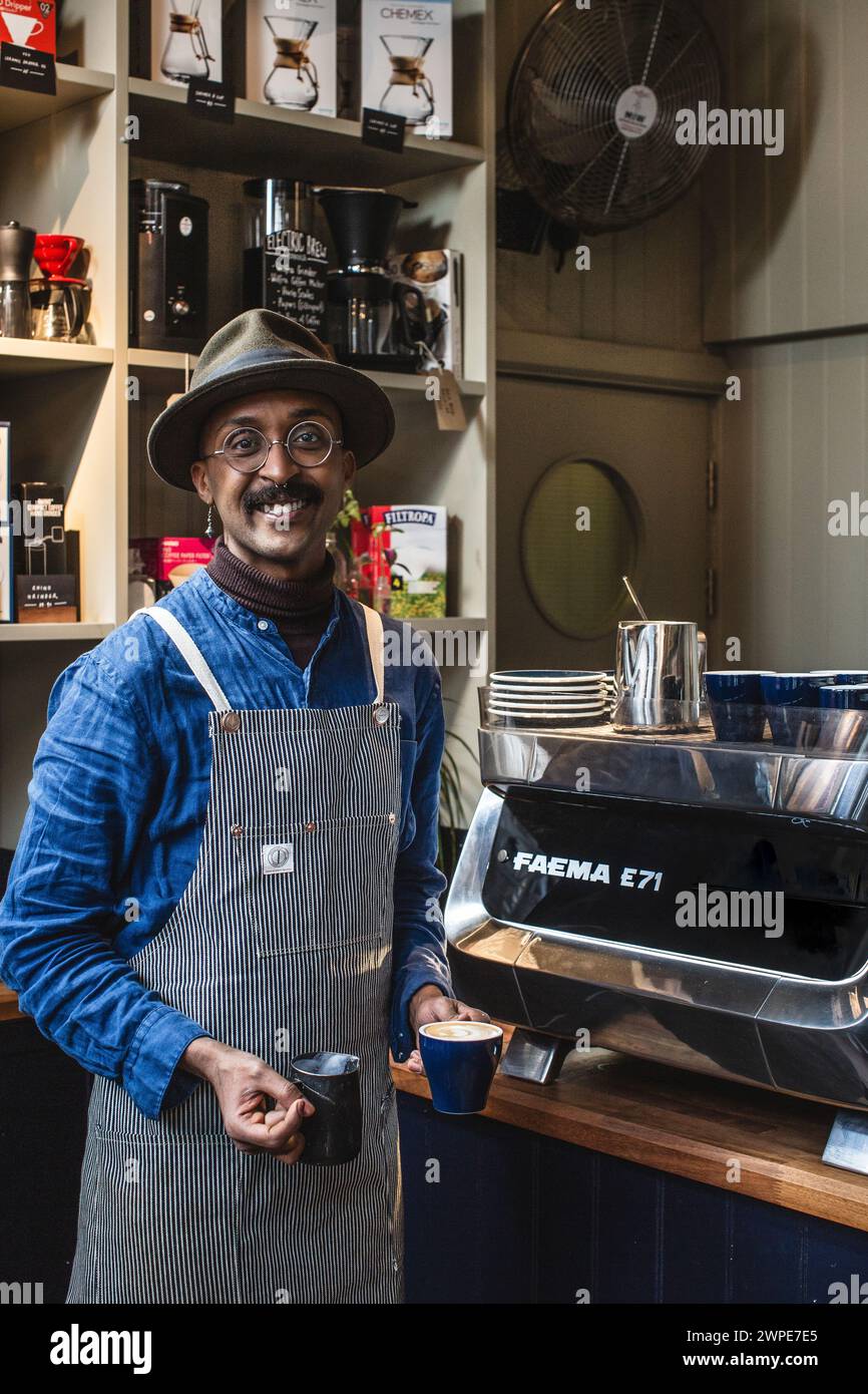 barista africano nero con grembiule per preparare caffè bianco piatto in caffetteria Foto Stock