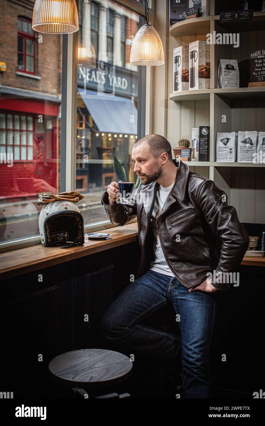 Un uomo che beve un caffè che guarda fuori da una finestra in un bar a Londra, indossa una giacca di pelle. Foto Stock