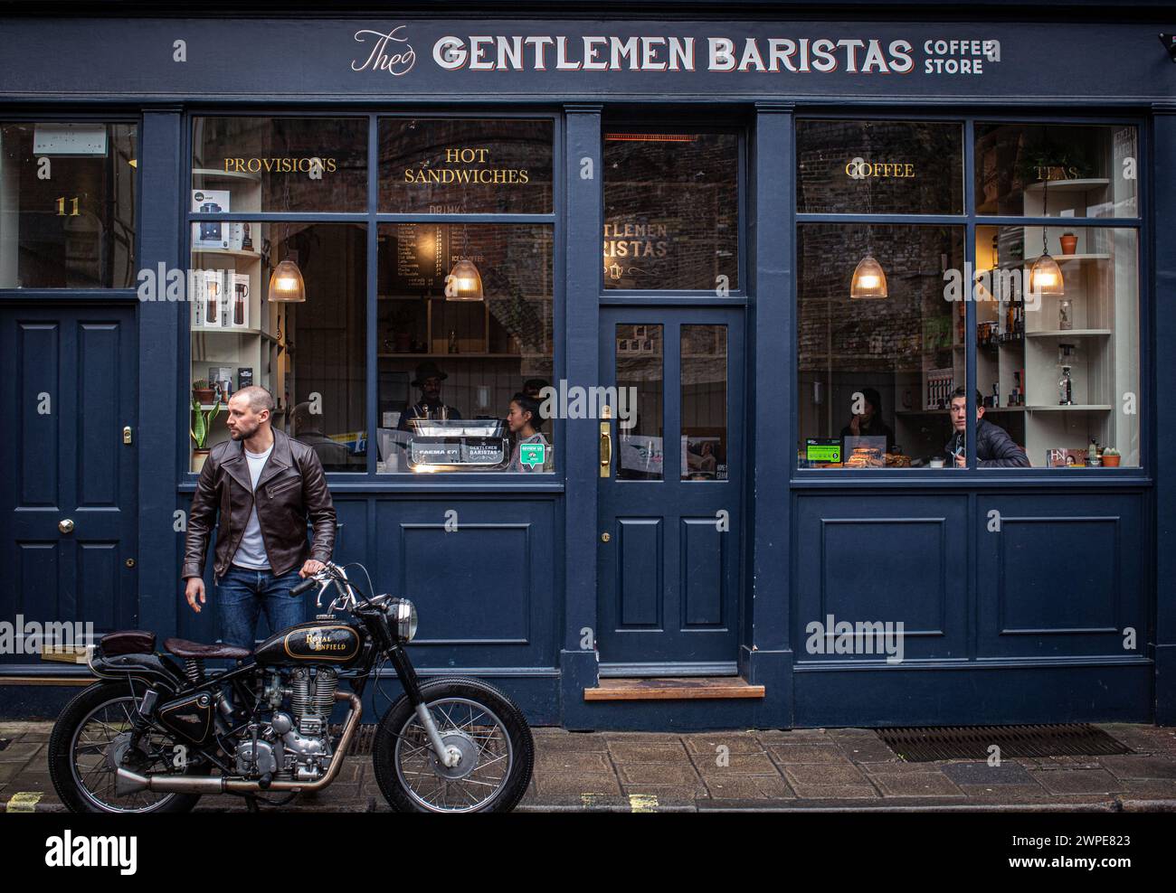 Un giovane uomo che indossa una giacca di pelle si prende una pausa caffè in un caffè a Londra con una classica moto parcheggiata di fronte a un bar Foto Stock