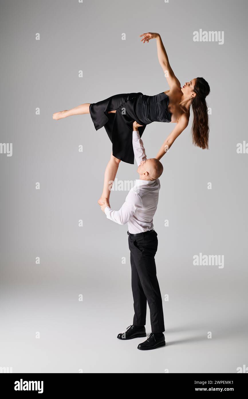 uomo forte che solleva una donna bruna flessibile in abito nero mentre si esibisce ballando su sfondo grigio Foto Stock