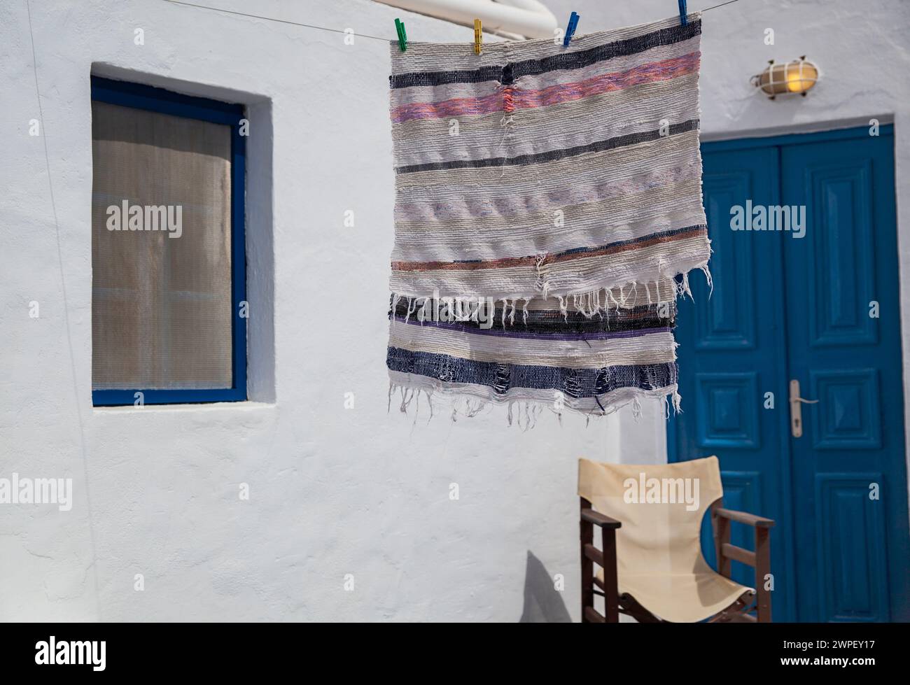 Un tessuto intrecciato a mano è appeso ad asciugare su una parete bianca di una casa greca, accanto a una porta blu, catturando l'essenza della vita dell'isola. Foto Stock