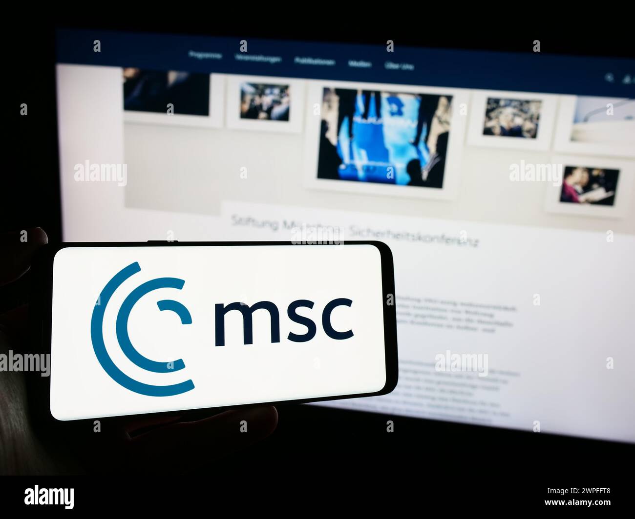 Persona che possiede un telefono cellulare con il logo dell'evento annuale Munich Security Conference (MSC) di fronte alla pagina Web. Mettere a fuoco il display del telefono. Foto Stock