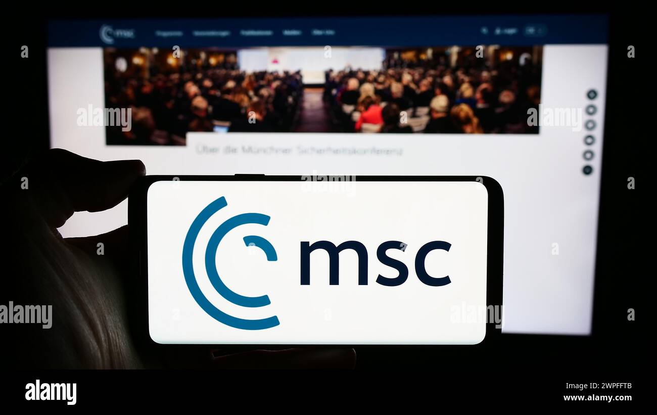 Persona che detiene un cellulare con il logo dell'evento annuale Munich Security Conference (MSC) di fronte alla pagina web. Mettere a fuoco il display del telefono. Foto Stock