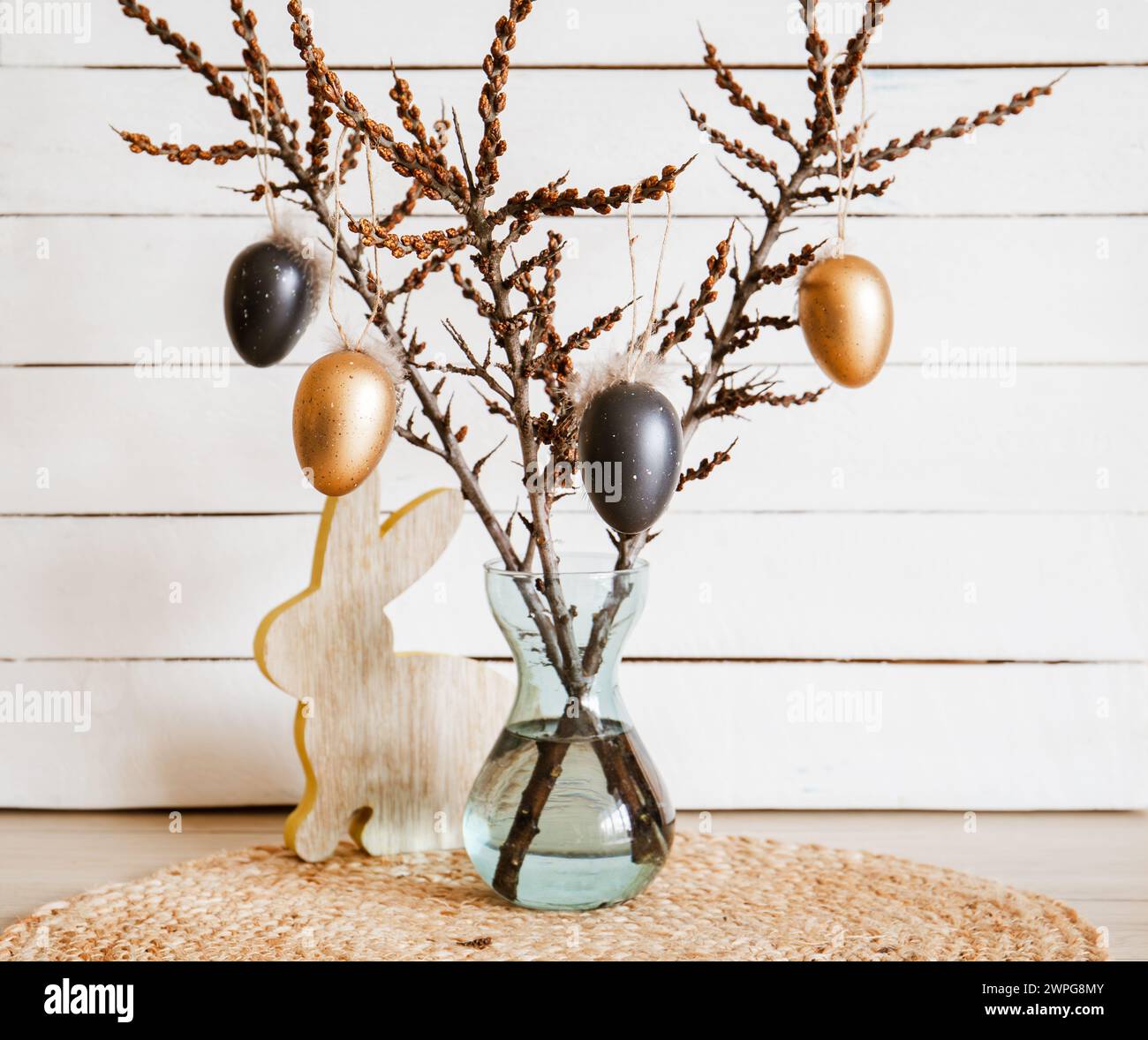 Rami d'albero in vaso con uova di Pasqua nere e di bronzo appese su spago, sfondo bianco rustico in legno. Decorazioni casalinghe minimali. Foto Stock