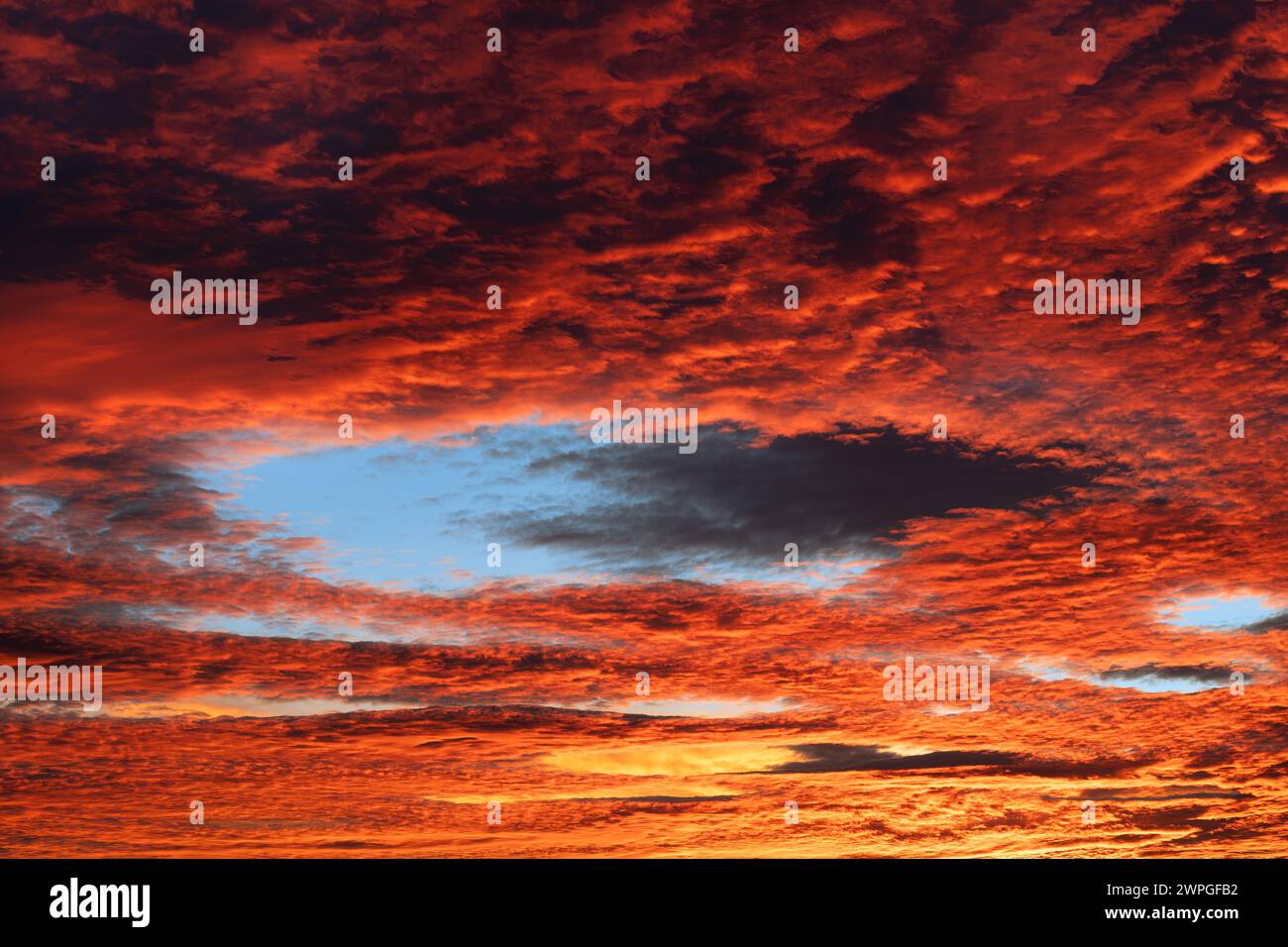 Paesaggio nuvoloso rosso con buco blu nelle nuvole cirrocumulative al crepuscolo serale. Un bellissimo bagliore di luce sul cielo arancione al tramonto con un'insolita cavum Foto Stock