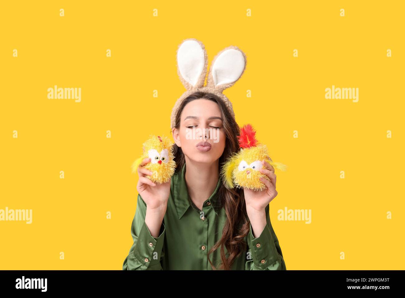 Giovane donna con le orecchie di coniglio pasquale fascia con polli giocattolo che soffiano baci su sfondo giallo Foto Stock