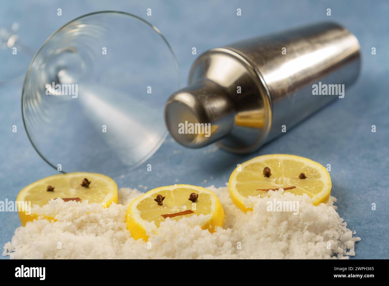 fette di limone con chiodi di garofano e cannella che formano una faccia sorridente su una pila di sale accanto a uno shaker per cocktail e un bicchiere di cristallo con sfondo blu Foto Stock