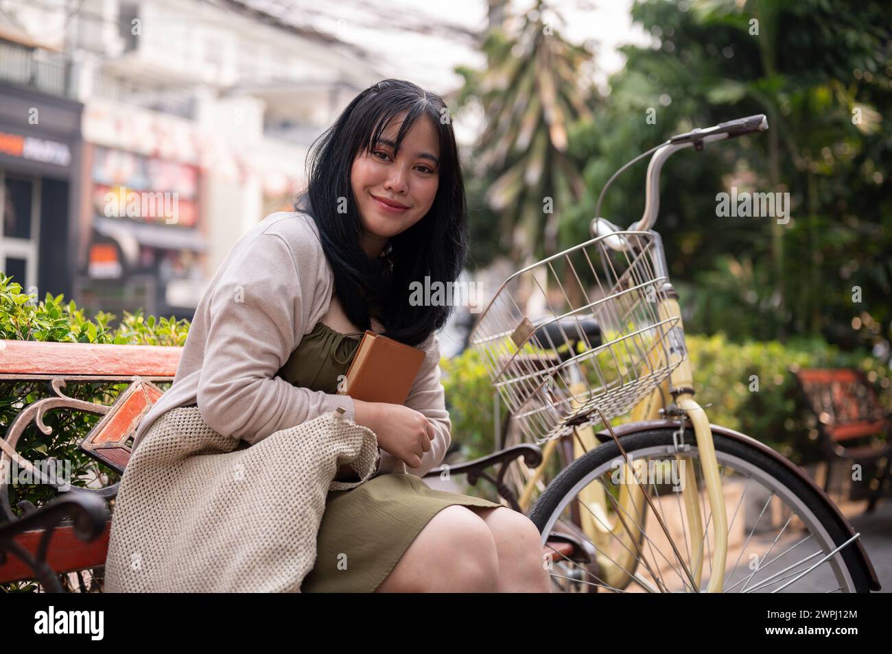Una giovane donna asiatica attraente e felice è seduta su una panchina all'aperto in città con la sua bici, rilassandosi in un parco pubblico cittadino. persone e stile di vita Foto Stock