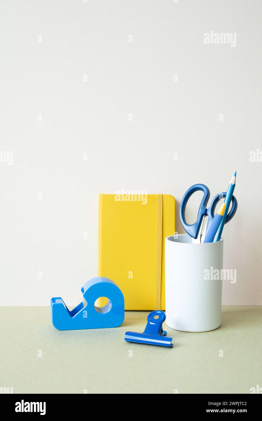 Quaderno giallo, matita blu, penna, forbici, clip, dispenser di nastro sulla scrivania. sfondo bianco avorio. cancelleria per ufficio Foto Stock