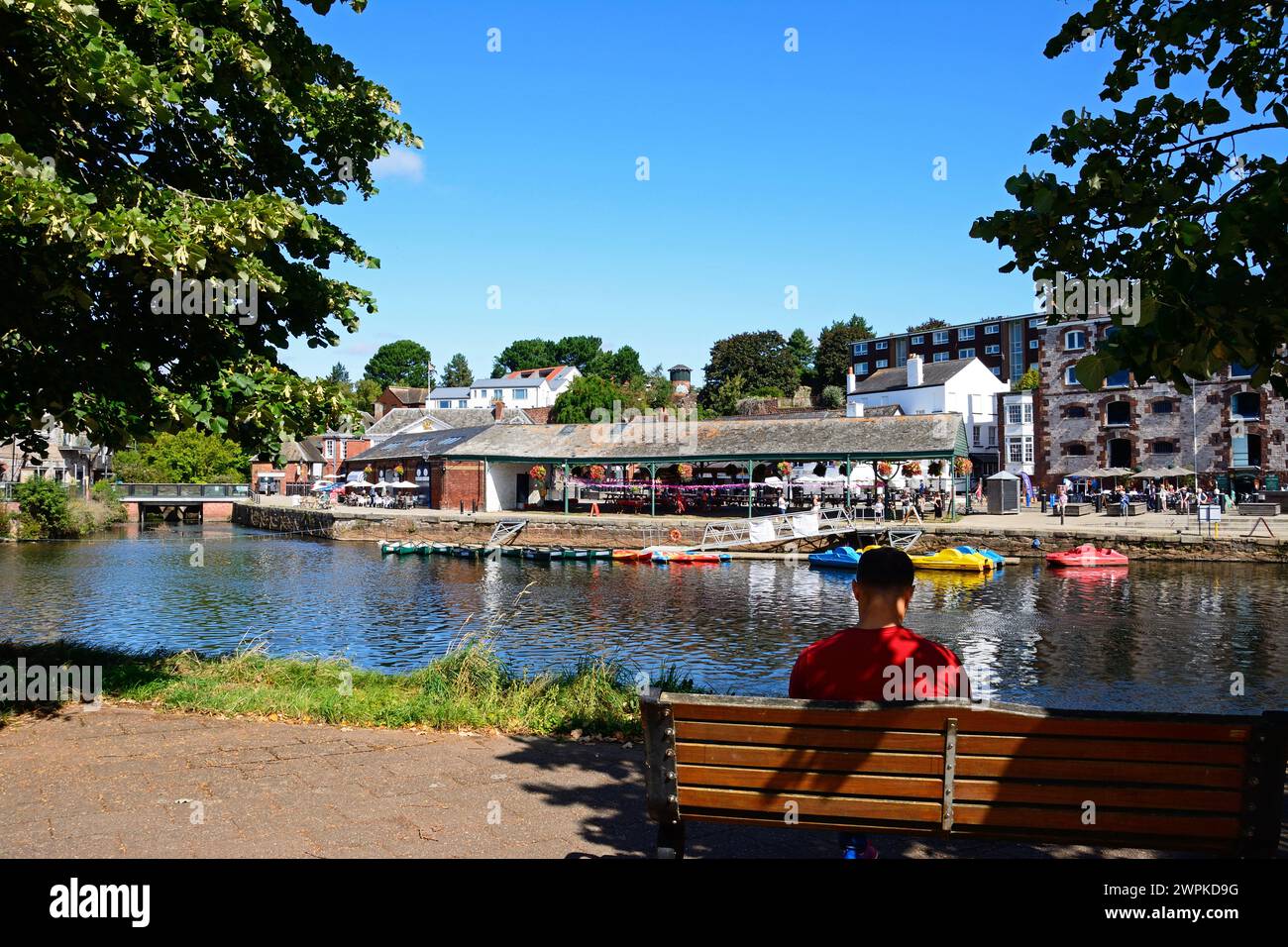 Uomo seduto su una panchina con vista sul fiume exe verso i negozi e i ristoranti lungo East Quay, Exeter, Devon, Regno Unito, Europa. Foto Stock