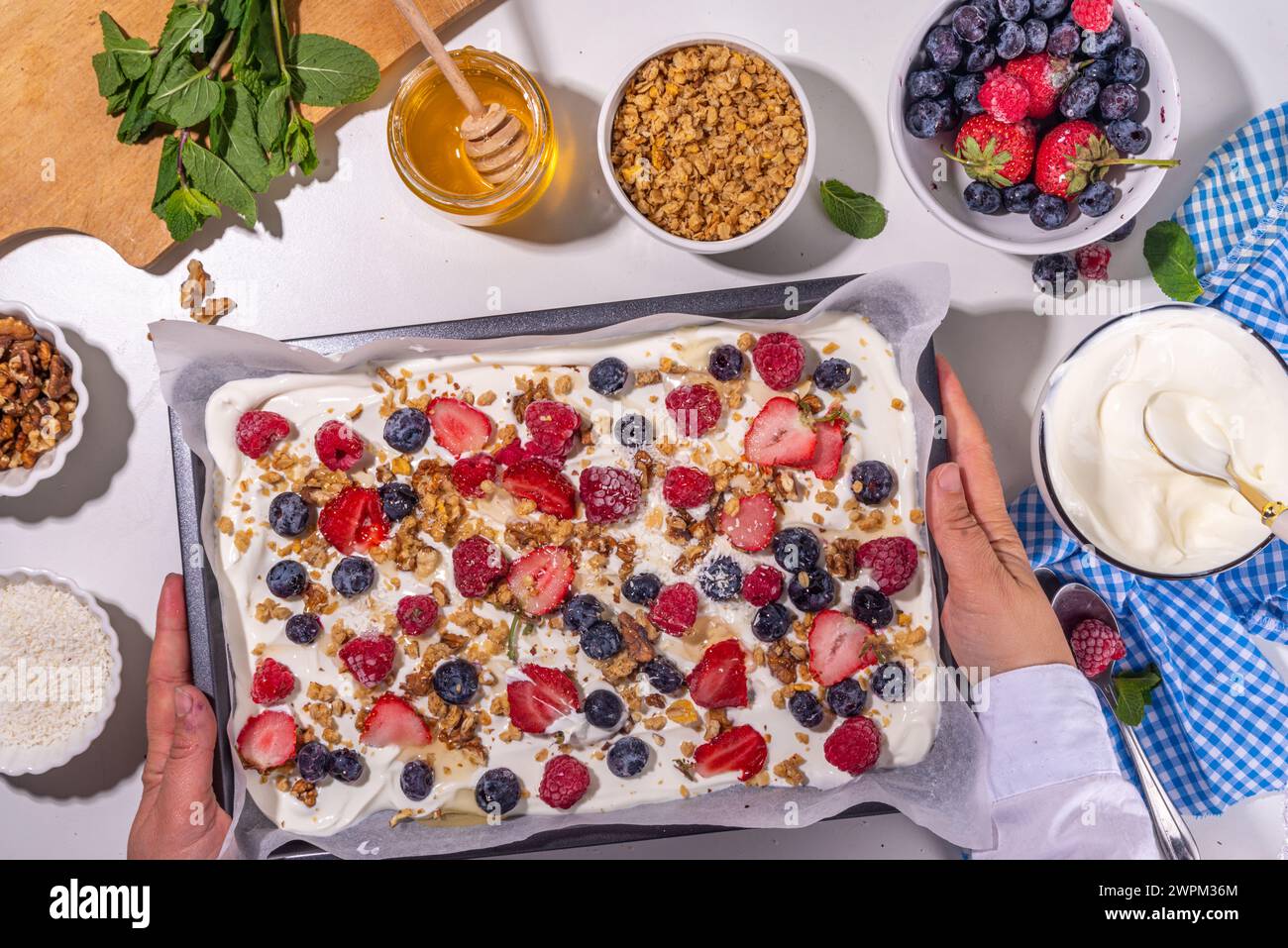 Preparazione della cottura con sfondo di corteccia di yogurt congelato con yogurt greco, bacche fresche, muesli, miele, salsa al cioccolato, con vassoio da forno deco. Vista dall'alto della donna Foto Stock