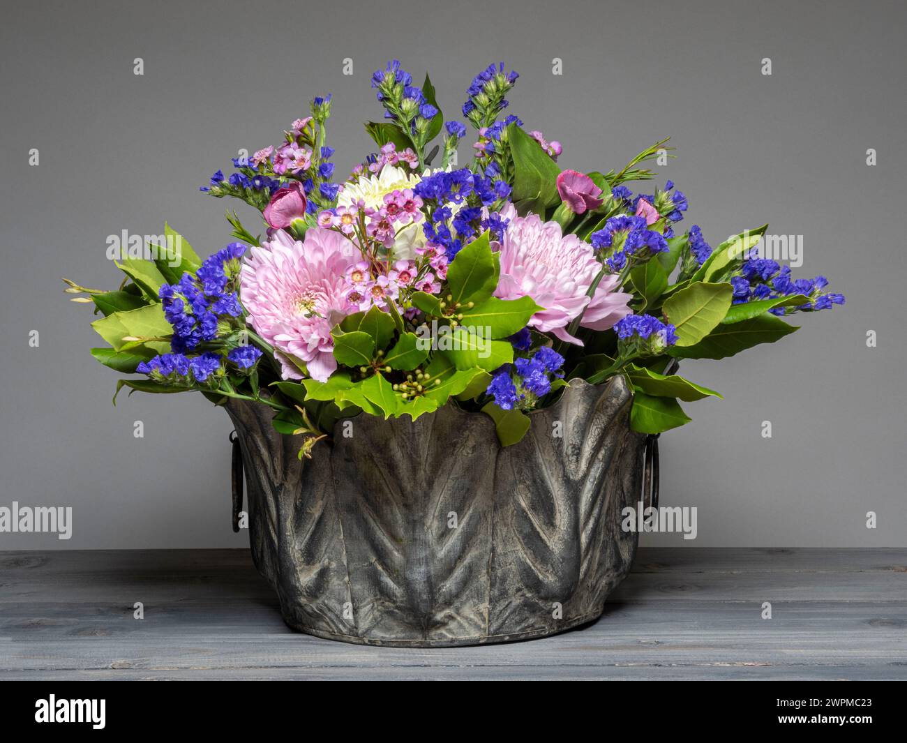 Fiori rosa e viola disposti con foglie di alloro in una piantatrice di cavoli galvanizzata, vista su uno sfondo grigio Foto Stock