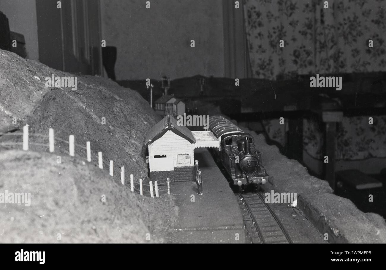 anni '1950, storico, sala interna di casa, un modello di ferrovia, immagine mostra un modello di treno a vapore su un binario ferroviario in una piattaforma della stazione. Foto Stock