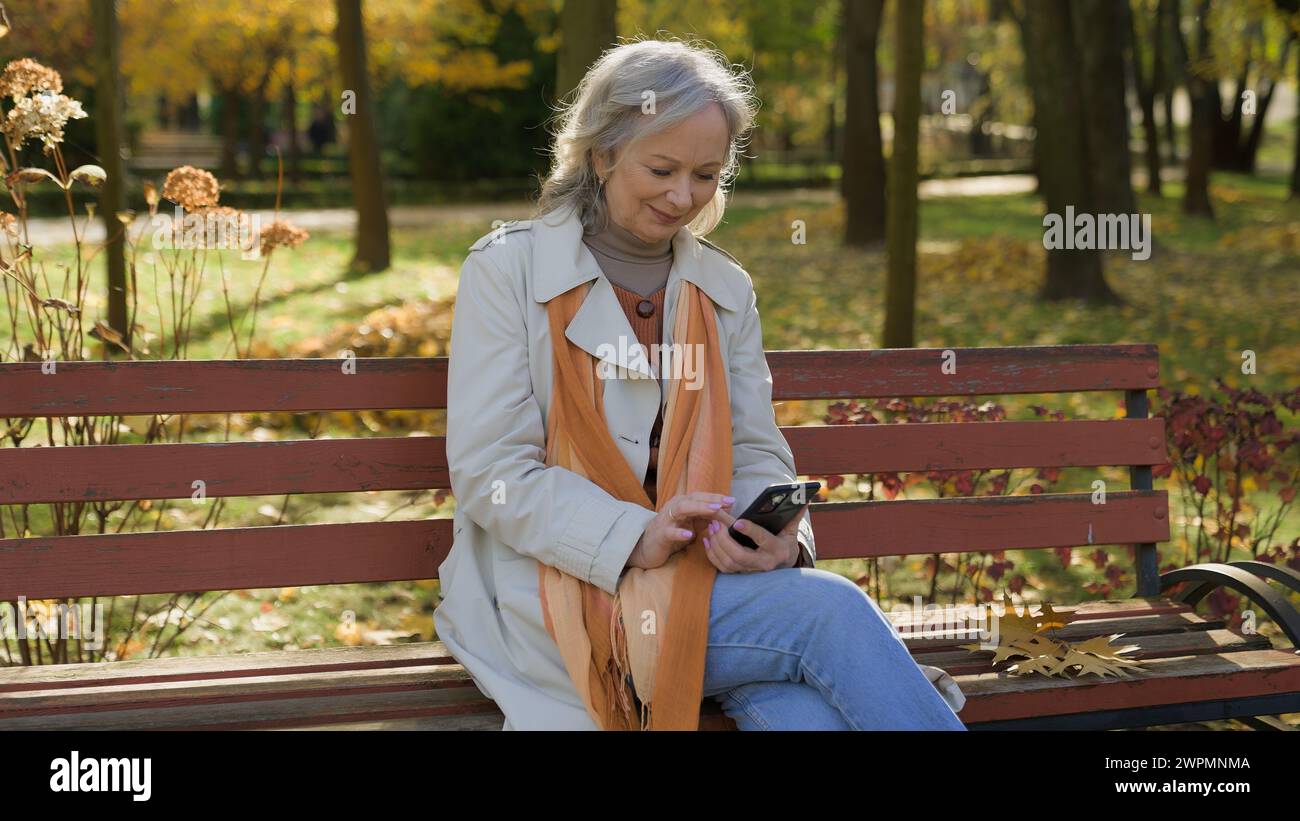 Felice dai capelli grigi bella donna matura che usa lo smartphone online nel parco autunnale donna caucasica che utilizza tecnologie Internet al di fuori della signora anziana Foto Stock