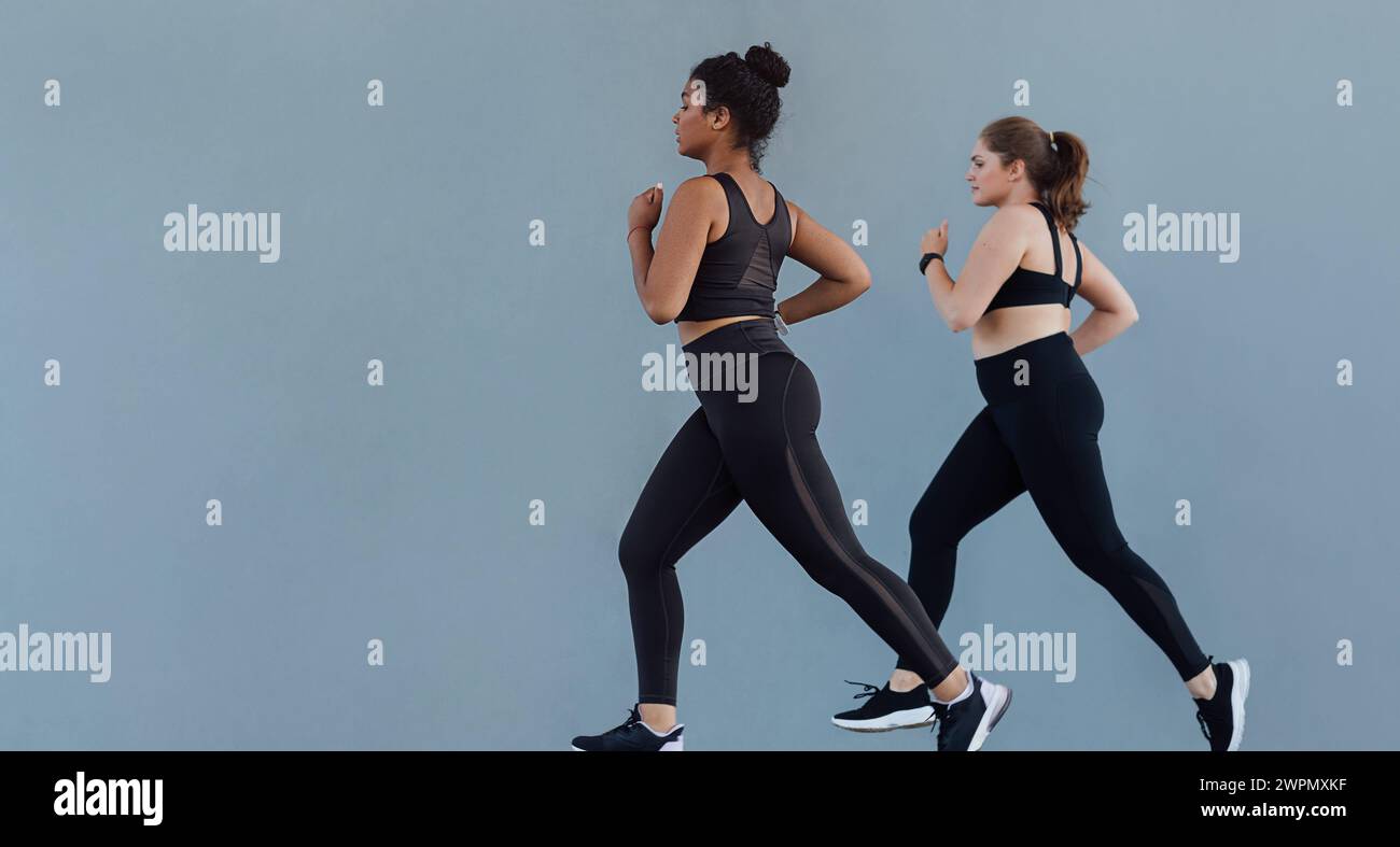 Lunghezza completa di due grandi donne che corrono sincronizzate su una parete grigia. Due donne in palestra indossano jogging all'aperto su un muro. Lunghezza completa di due più Foto Stock