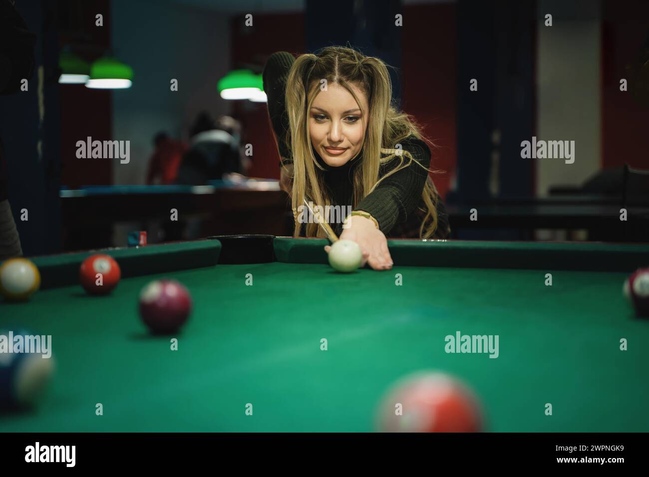 Una donna con lunghi capelli biondi ondulati, vestita con un top nero, è concentrata su un tiro in una partita di biliardo a biliardo - giocatore di tavolo da biliardo - tempo libero Foto Stock